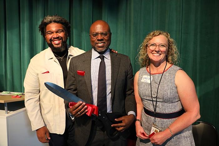 three people are shown from just below the waist. the one in the center holds giant scissors